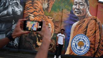 Seorang pria berpose untuk foto didepan lukisan pemilik klub Leicester City Srivaddhanaprabha dekat Universitas De Montfort, Leicester, Inggris, Minggu (28/10). [AFP/Ben STANSALL]