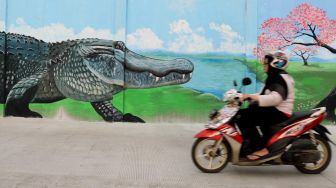 Pengendara motor melewati mural bergambar Buaya di bantaran kali Opak, Jakarta Utara, Selasa (11/02). [Suara.com/Alfian Winanto]