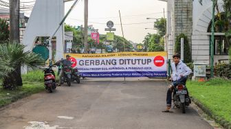 Pengendara motor berdiri di depan spanduk pengumuman berisi karantina wilayah perumahan elit Kemang Pratama, Bekasi, Jawa Barat, Rabu (1/4). [Suara.com/Alfian Winanto]