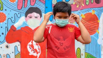 Seorang anak memakai masker didepan mural bertemakan virus Corona di Cipayung, Depok, Jawa Barat, Rabu (15/4). [Suara.com/Alfian Winanto]