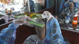 Tukang cukur menggunakan APD (Alat Pelindung Diri) saat mencukur pelanggan di kawasan Pondok Kelapa, Jakarta Timur, Selasa (5/5). Suara.com/Angga Budhiyanto]