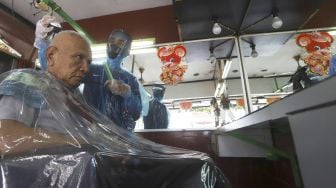 Tukang cukur menggunakan APD (Alat Pelindung Diri) saat mencukur pelanggan di kawasan Pondok Kelapa, Jakarta Timur, Selasa (5/5). Suara.com/Angga Budhiyanto]