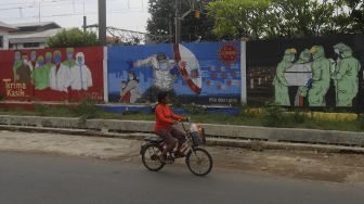 Pesepeda melintas di depan mural tentang pandemi COVID-19 di Bukit Duri, Jakarta, Selasa (20/10/2020). [Suara.com/Angga Budhiyanto]