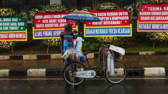 Pedagang kopi keliling melayani pembeli didepan karangan bunga terkait kasus korupsi Jiwasraya didepan Kejaksaan Agung RI, Jakarta Selatan, Selasa (27/10). [Suara.com/Alfian Winanto]