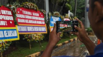 Seorang warga mengabadikan dengan smartphonenya karangan bunga terkait kasus korupsi Jiwasraya didepan Kejaksaan Agung RI, Jakarta Selatan, Selasa (27/10). [Suara.com/Alfian Winanto]