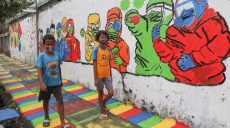 Anak - anak melintas di depan mural edukasi Covid-19 di Kampung Tangguh Jaya, Penggilingan, Cakung, Jakarta Timur, Minggu (17/1/2021). [Suara.com/Alfian Winanto]