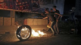 Pengunjuk rasa Palestina menggulingkan ban yang terbakar di tengah bentrokan dengan pasukan keamanan Israel di pos pemeriksaan Qalandia antara kota Ramallah di Tepi Barat dan Yerusalem, pada (8/5/2021). [ABBAS MOMANI / AFP]