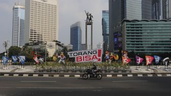 Pengendara sepeda motor melintas di dekat maskot dan slogan Pekan Olahraga Nasional (PON) XX Papua di Bundaran Hotel Indonesia, Jakarta, Jumat (16/7/2021). [Suara.com/Angga Budhiyanto]