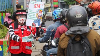 Angggota Komunitas Aku Badut Indonesia (ABI) membagikan masker kepada pengendara motor saat melakukan aksi kampanye untuk mengajak masyarakat melakukan vaksinasi dan mematuhi protokol kesehatan di Kawasan Juanda, Depok, Jawa Barat, Jumat (23/7/2021). [Suara.com/Alfian Winanto]