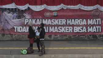 Warga berdiri di depan spanduk berisi imbauan untuk memakai masker yang terpasang di Jalan Gatot Subroto, Jakarta, Senin (23/8/2021). [Suara.com/Angga Budhiyanto]