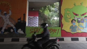 Pengendara sepeda motor melintas di dekat mural bertema COVID-19 di kolong Jalan Tol Lingkar Luar Jakarta, Cipayung, Jakarta Timur, Jumat (3/9/2021). [Suara.com/Angga Budhiyanto]