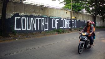 Pengendara sepeda motor tengah melintas di mural bertuliskan “ Country of Jokes” di Tangerang, Jumat (3/9).  ( Suara.com/Hilal Rauda Fiqry) 