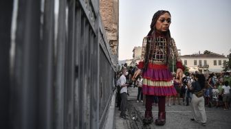 "Little Amal", boneka raksasa yang menggambarkan seorang gadis pengungsi muda berdiri di dekat pagar saat diarak di beberapa jalan di Athena, Yunani, pada (2/9/2021). [Louisa GOULIAMAKI / AFP]