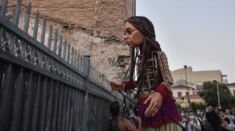 "Little Amal", boneka raksasa yang menggambarkan seorang gadis pengungsi muda berdiri di dekat pagar saat diarak di beberapa jalan di Athena, Yunani, pada (2/9/2021). [Louisa GOULIAMAKI / AFP]