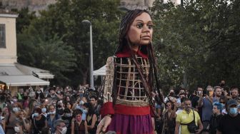 Warga berkumpul di sekitar "Little Amal", boneka raksasa yang menggambarkan seorang gadis pengungsi muda saat diarak di beberapa jalan di Athena, Yunani, pada (2/9/2021). [Louisa GOULIAMAKI / AFP]