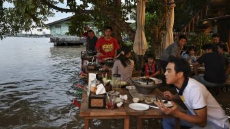 Pengunjung berdiri sambil menikmati makan malam di Kafe Antik Chaopraya, saat air banjir dari Sungai Chao Phraya mengalir ke restoran di provinsi Nonthaburi, Bangkok, Thailand, pada (7/10/2021). [Lillian SUWANRUMPHA / AFP]