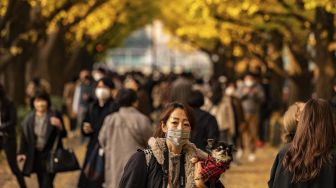 Seorang warga membawa anjingnya sambil berjalan di bawah pohon ginkgo dalam warna musim gugur di Taman Luar Kuil Meiji, Tokyo, pada (18/11/2021). [PHILIP FONG / AFP]