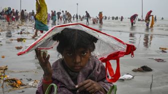 Seorang anak menutupi kepalanya dengan kantong plastik untuk berlindung dari hujan dengan  latar belakang peziarah Hindu berkumpul di tepi Gangga dan Teluk Benggala selama Gangasagar Mela pada kesempatan Makar Sankranti di Pulau Sagar, India, pada (14/1/2022). [DIBYANGSHU SARKAR / AFP]