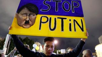 Seorang demonstran mengangkat tanda yang menunjukkan bendera Ukraina dengan wajah Adolf Hitler Nazi Jerman dan teks dalam bahasa Inggris bertuliskan "Hentikan Putin" selama protes terhadap operasi militer Rusia di Ukraina, di depan kedutaan Rusia, Tel Aviv, Israel, Sabtu (26/2/2022). [JACK GUEZ / AFP]