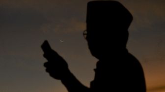 Seorang jamaah Tarekat Syattariyah mengecek ponselnya dengan latar bulan sabit, saat melakukan tradisi maniliak bulan (hilal), di Pantai Ulakan, Kabupaten Padangpariaman, Sumatera Barat, Minggu (03/4/2022). [ANTARA FOTO/Iggoy el Fitra/YU]