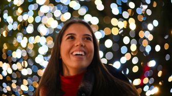 Pengunjung berpose di bawah instalasi cahaya saat perayaan natal Kew atau &quot;Christmas at Kew&quot; di Kew Gardens, barat daya London, Inggris, Selasa (15/11/2022). [Daniel LEAL / AFP]