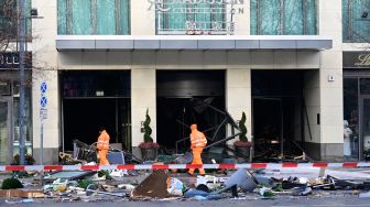 Pekerja departemen kebersihan kota berjalan melewati puing-puing yang menutupi jalan di depan hotel Radisson Blu setelah akuarium besar yang terletak di lobi hotel meledak di Berlin, Jerman, Jumat (16/12/2022). [John MACDOUGALL / AFP]