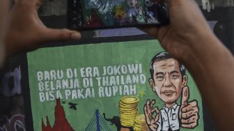 Warga memotret mural yang bertuliskan “Baru di era Jokowi belanja di Thailand bisa pakai rupiah” di Kabupaten Ciamis, Jawa Barat, Jumat (30/12/2022). [ANTARA FOTO/Adeng Bustomi].