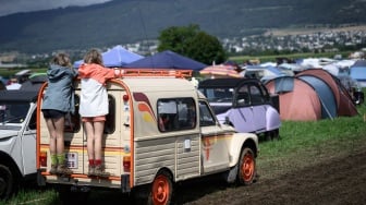 Dua gadis muda terlihat bergelantungan di belakang mobil saat pertemuan Dunia ke-24 Citroen 2CV Friends di dekat Delemont, Swiss, Rabu (26/7/2023). [GABRIEL MONNET/AFP]