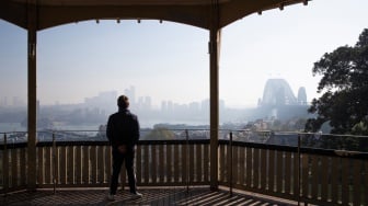 Seorang pria melihat keluar dari balkonnya ke arah cakrawala yang diselimuti kabut asap di Sydney, Australia, Rabu (13/9/2023). [Steve CHRISTO / AFP]