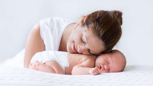 Young mother hugging her newborn child. Mom nursing baby. Woman and new born boy relax in a white bedroom. Family at home. Love, trust and tenderness concept. Bedding and textile for nursery