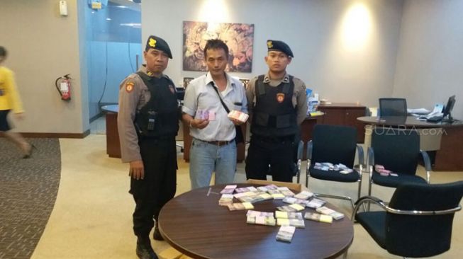 Mujiono mengenakan baju putih diapit petugas kepolisian menunjukkan uang mainan saat di Bank BCA Jalan Pangeran Diponegoro, Tulungagung. [Suara.com/Ali Achmad]