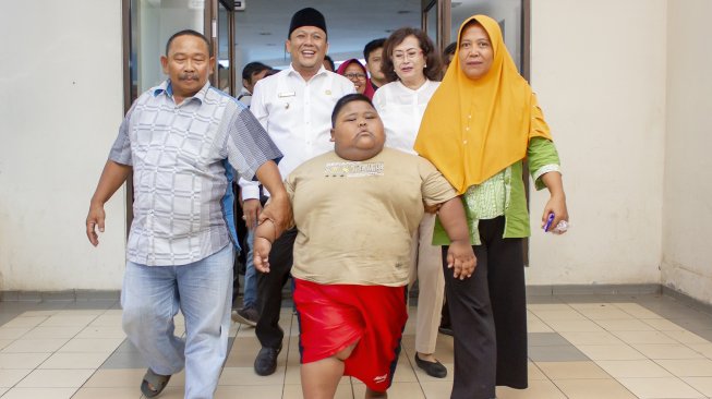 Satia Putra (7) penyandang obesitas dengan berat badan mencapai 101 kg didampingi Wakil Bupati Karawang Ahmad Zamaksyari (kedua kiri) bersama kedua orangtuanya berjalan menuju lokasi pemeriksaan medis di Rumah Sakit Umum Daerah (RSUD) Karawang, Karawang, Jawa Barat, Rabu (3/7). [ANTARA FOTO/M Ibnu Chazar]