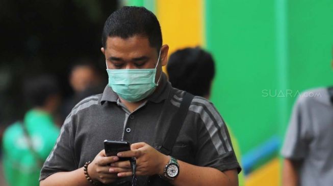 Warga melihat smartphonenya dengan menggunakan masker di kawasan Pasar Pramuka, Jakarta, Selasa (11/2). [Suara.com/Alfian Winanto]