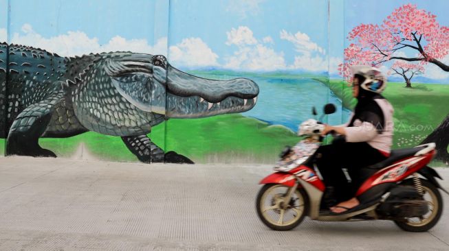 Pengendara motor melewati mural bergambar Buaya di bantaran kali Opak, Jakarta Utara, Selasa (11/02). [Suara.com/Alfian Winanto]