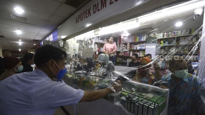 Pekerja melayani pembeli dari balik plastik pembatas di salah satu apotek di kawasan Pasar Baru, Jakarta, Jumat (3/4). [Suara.com/Angga Budhiyanto]