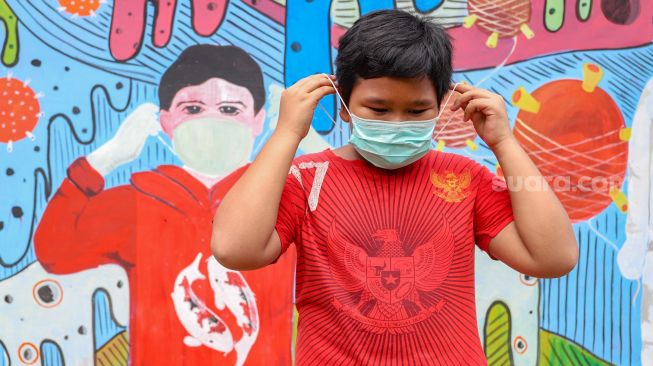 Seorang anak memakai masker didepan mural bertemakan virus Corona di Cipayung, Depok, Jawa Barat, Rabu (15/4). [Suara.com/Alfian Winanto]