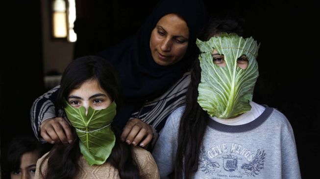 Seorang ibu memakaikan anak-anaknya masker dari daun kubis di Beit Lahia, Jalur Gaza, Palestina, Kamis (16/4/2020).  (AFP/Mohamed Abed)