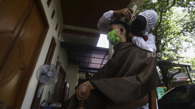 Seorang tukang cukur menggunakan Alat Pelindung Diri (APD) mencukur rambut di halaman rumah pelanggan di kawasan Kelapa Dua, Depok, Jawa Barat, Rabu  (29/4). [Suara.com/Angga Budhiyanto]