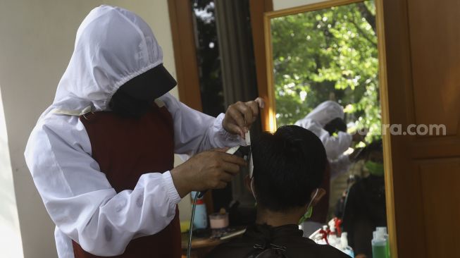 Seorang tukang cukur menggunakan Alat Pelindung Diri (APD) bersiap memotong rambut di halaman rumah pelanggan di kawasan Kelapa Dua, Depok, Jawa Barat, Rabu  (29/4). [Suara.com/Angga Budhiyanto]