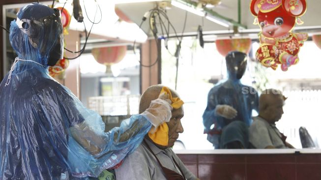 Tukang cukur menggunakan APD (Alat Pelindung Diri) saat merapihkan rambut pelanggan di kawasan Pondok Kelapa, Jakarta Timur, Selasa (5/5). Suara.com/Angga Budhiyanto]