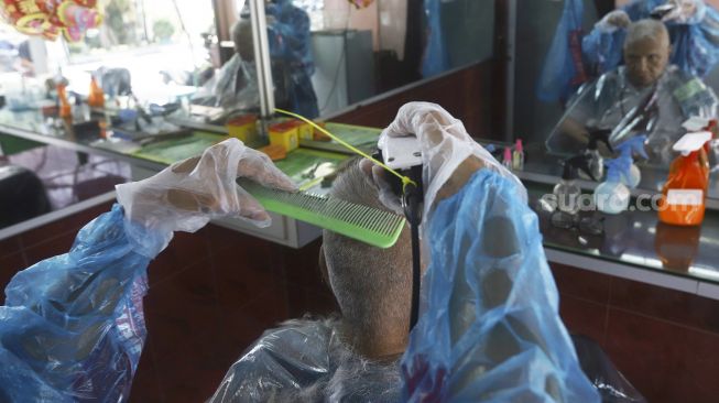 Tukang cukur menggunakan APD (Alat Pelindung Diri) saat mencukur pelanggan di kawasan Pondok Kelapa, Jakarta Timur, Selasa (5/5). Suara.com/Angga Budhiyanto]