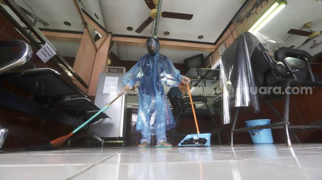 Tukang cukur menggunakan APD (Alat Pelindung Diri) membersihkan tempat cukur usai mencukur pelanggan di kawasan Pondok Kelapa, Jakarta Timur, Selasa (5/5). [Suara.com/Angga Budhiyanto]
