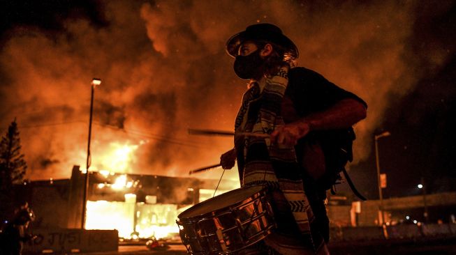 Seorang demonstran memainkan drum di depan gedung yang terbakar saat demonstrasi di Minneapolis, Minnesota, Jumat (29/5). [Chandan Khanna/ AFP]