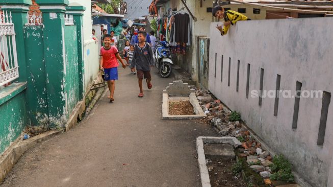 Sejumlah bocah melintasi kuburan yang ada di jalanan rumah warga di Pisangan, Jakarta Timur, Selasa (16/5). [Suara.com/Alfian Winanto]