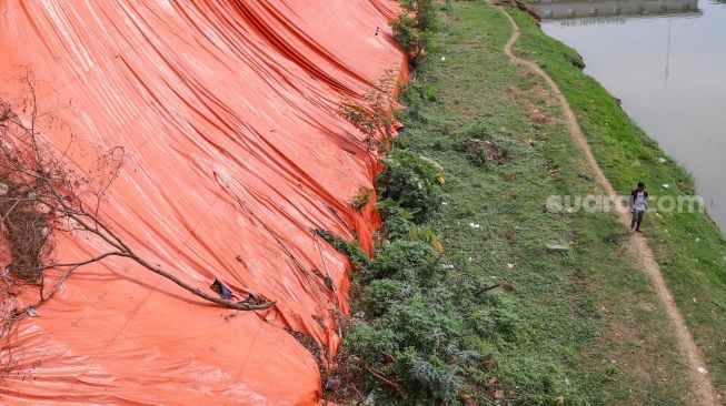 Warga melintas dibawah area longsor di pinggir Banjir Kanal Timur (BKT), Malaka Sari, Jakarta Timur, Jumat (19/6). [Suara.com/Alfian Winanto]