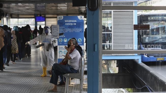 Anggota komunitas pecinta Transjakarta mengenakan baju hazmat dengan membawa poster berisi informasi jumlah kasus positif di Jakarta saat melakukan sosialisasi protokol kesehatan di Halte Transjakarta Harmoni, Jakarta, Jumat (17/7/2020). [Suara.com/Angga Budhiyanto]