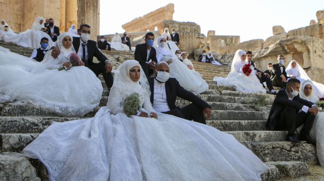 Sejumlah pasangan pengantin mengenakan masker wajah duduk di tangga selama acara pernikahan bersama di tengah pandemi Covid-19 di Kuil Bacchus di situs bersejarah Baalbek di Lembah Bekaa, Lebanon Timur, Jumat (24/7/2020). [AFP]