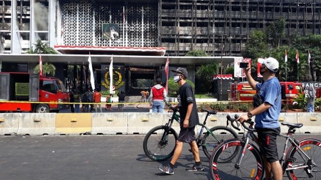 Pesepeda berhenti dan berfoto di depan gedung Kejagung yang terbakar, Minggu (23/8/2020). (Suara.com/Novian Ardiansyah)