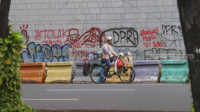 Pedagang kopi keliling melintas didepan dinding Kedutaan Besar Amerika Serikat yang masih dipenuhi coretan aksi demo tolak Omnibus Law Kamis (8/10) lalu di Jalan Merdeka Selatan, Jakarta Pusat, Sabtu (10/10). [Suara.com/Alfian Winanto]