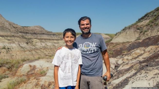 Nathan Krushkin dan ayahnya berpose di situs penggaloan fosil Hadrosaur berusia 69 juta tahun di Alberta, Kanada. [NATURE CONSERVANCY OF CANADA/BBC]